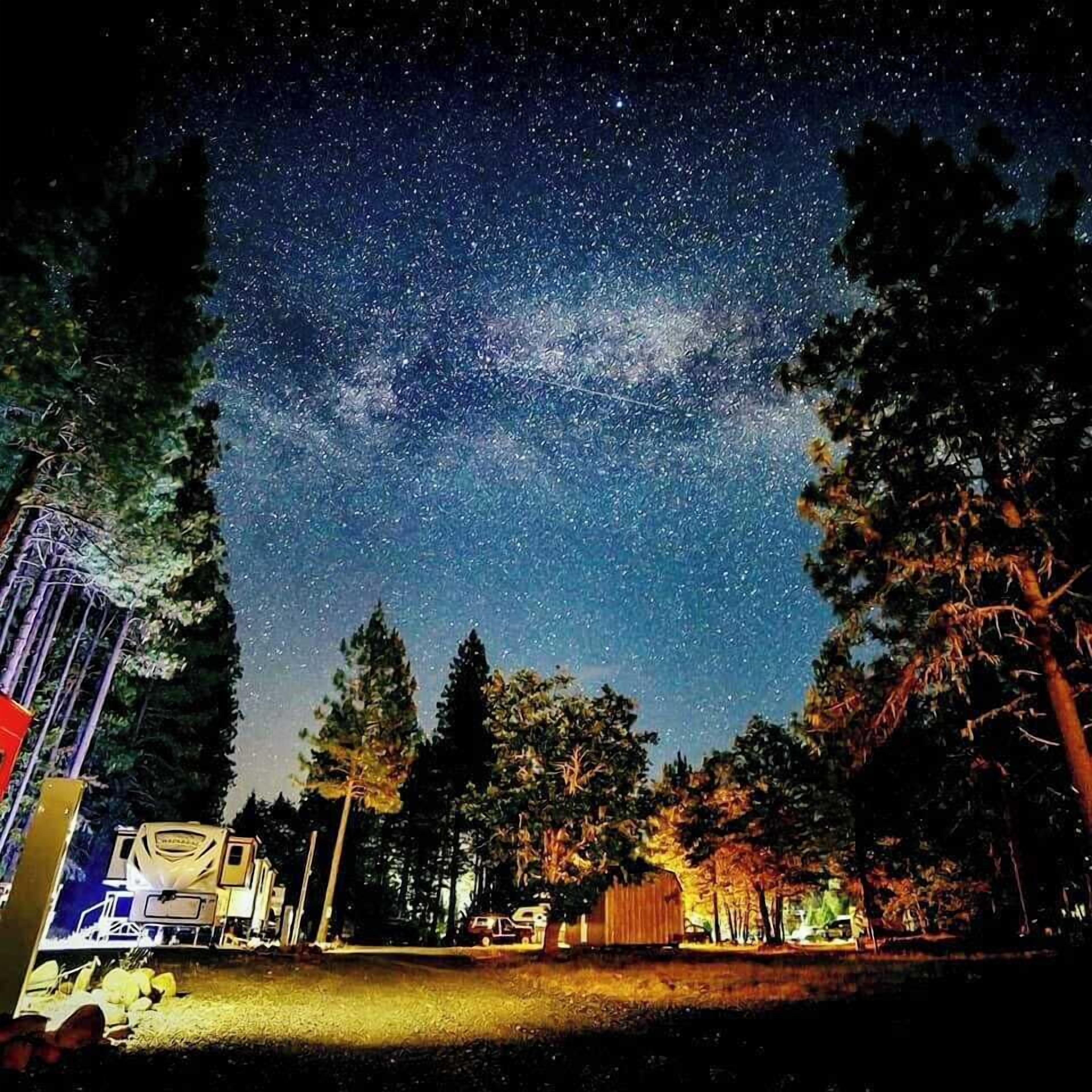 A campsite with a trailer and a vehicle, surrounded by trees under a starry night sky.