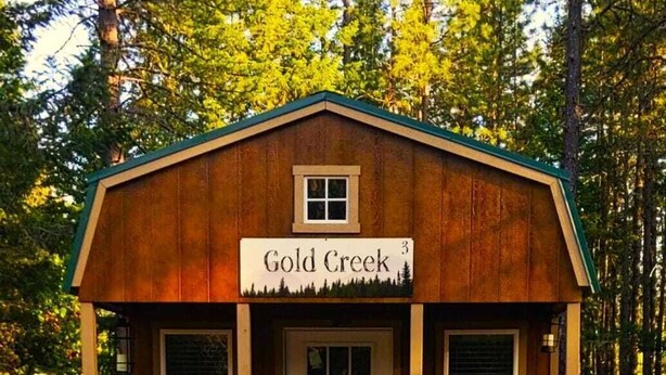 Eine kleine Holzhütte mit der Aufschrift "Gold Creek", einer Haustür, zwei Fenstern und zwei Adirondack-Stühlen davor.