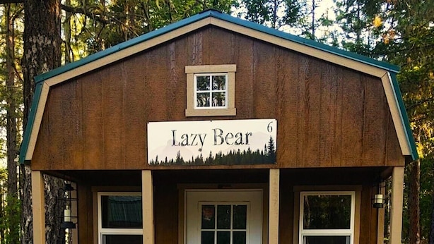 Een kleine houten blokhut met een bord waarop 'Lazy Bear' staat en twee Adirondack-stoelen ervoor.