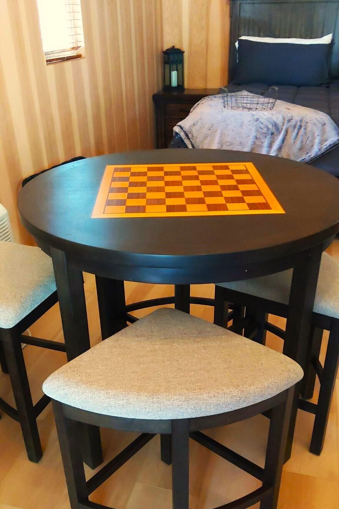 A round table with a chessboard, four chairs, a bed, and a nightstand with a lantern in a room with wooden walls.