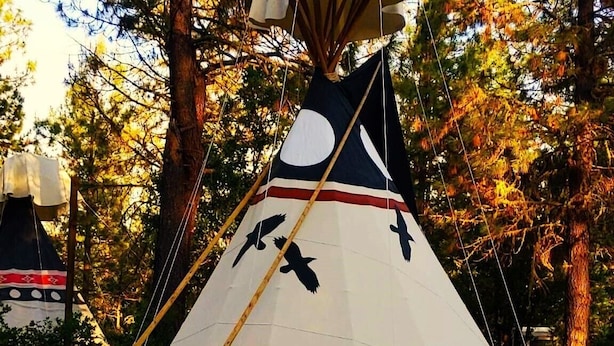 En stor tipi-telt med kegleformet tag, placeret på en træterrasse omgivet af træer.