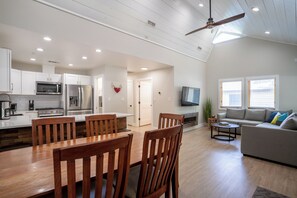 A spacious living area with a dining table, wooden chairs, a grey sofa, and a TV mounted on the wall.