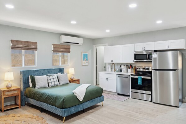 A bedroom with a bed, bedside tables, and a nightstand. There is a kitchen area with a refrigerator, microwave, oven, and cabinets.