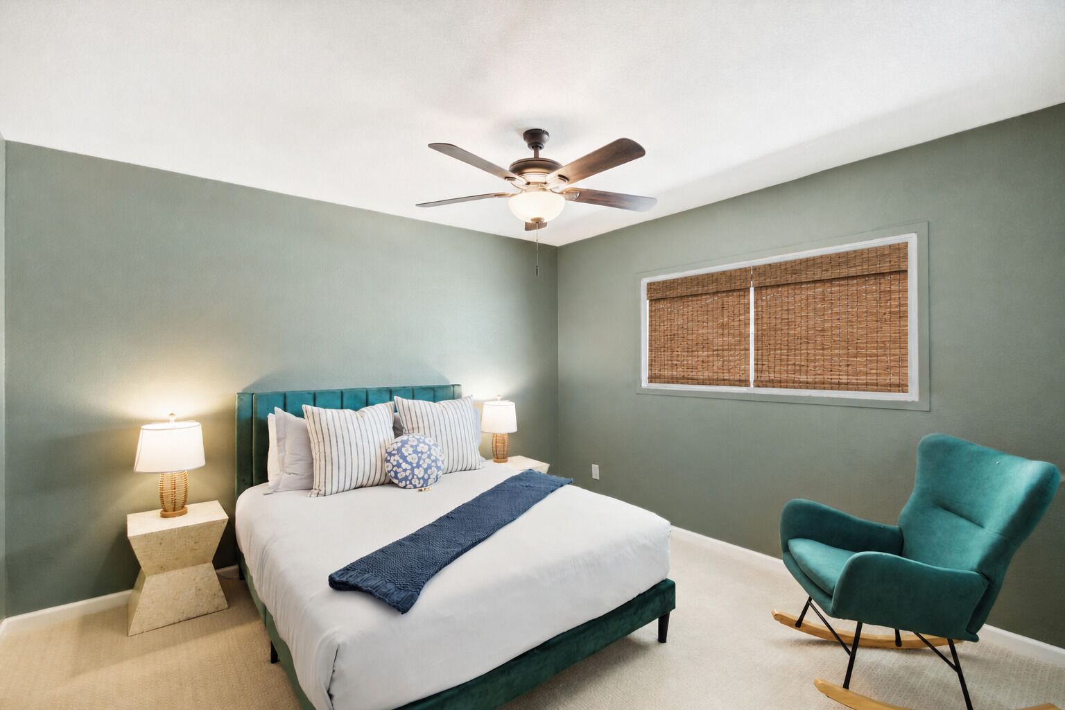 A bedroom with a bed, a ceiling fan, a window with blinds, a chair, and bedside lamps.
