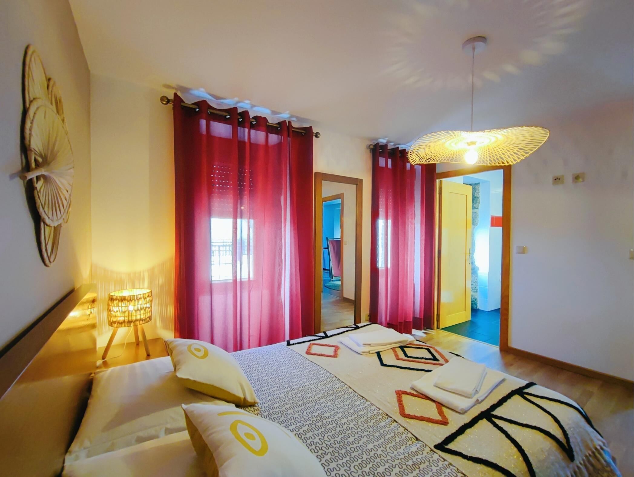 A bedroom with a bed, red curtains, a hanging light fixture, and a view of a hallway.