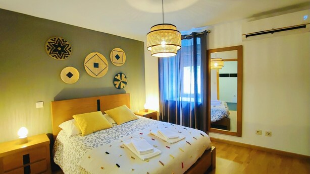 Una cama bien hecha con una colcha estampada y almohadas, una lámpara de noche, un espejo montado en la pared y decoración en la pared.