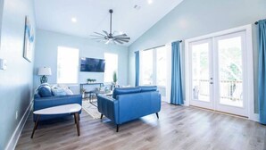 A spacious living room with a blue sofa, a coffee table, and a TV. There are large windows with curtains, a ceiling fan, and a wooden floor.