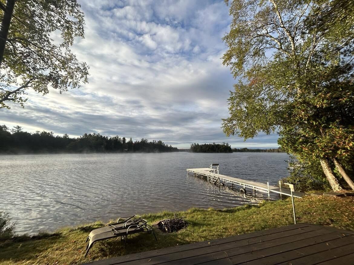 Une terrasse au bord du lac, avec un quai s’avançant dans une eau calme, entourée d’arbres et surplombée par un ciel nuageux.