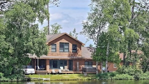 Een houten chalet met twee verdiepingen, een steiger en omringende bomen en water.