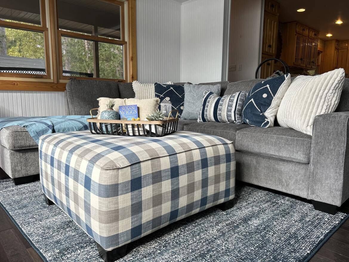 A cozy living room with a grey sectional sofa, a plaid ottoman, and a small wooden coffee table.
