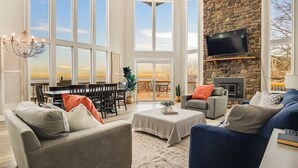 A spacious living room with large windows, a stone fireplace, and a television mounted on the wall. - Private Pool, Mountain Top Views & Home Theater (Hot Springs)