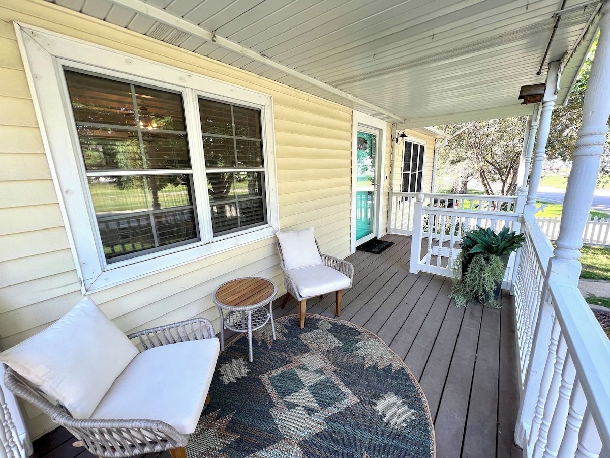 A covered porch with two white wicker chairs, a small round table, and a patterned rug.