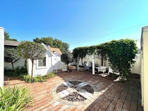 A wooden deck with a circular fire pit, surrounded by potted plants and a white fence. - Sunshine Cottage | Near Whitewater Park | Sleeps 10 (Norfolk)