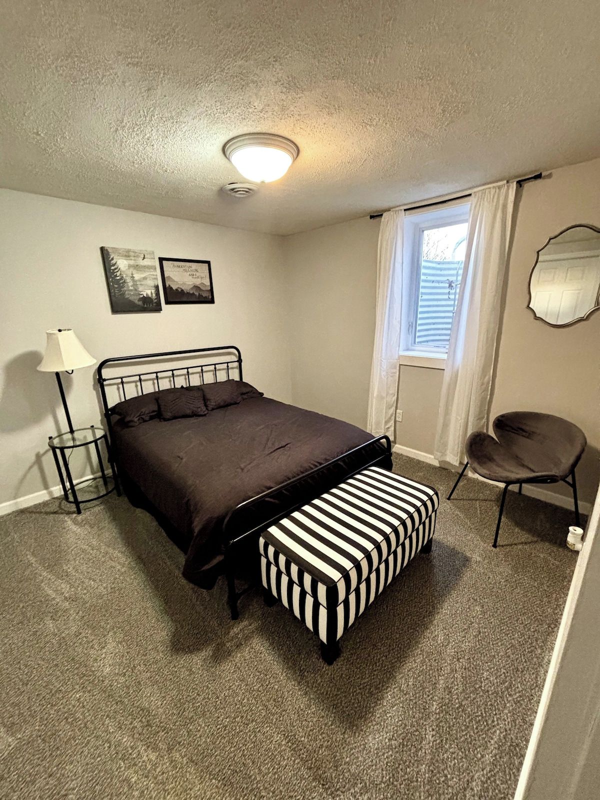 A bedroom with a bed, a bedside lamp, a window with curtains, a chair, and a small table.