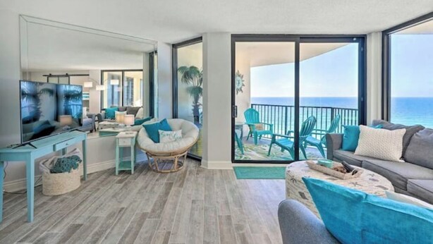 A modern living room with a large TV, a dining area, and a balcony with a view of the ocean.