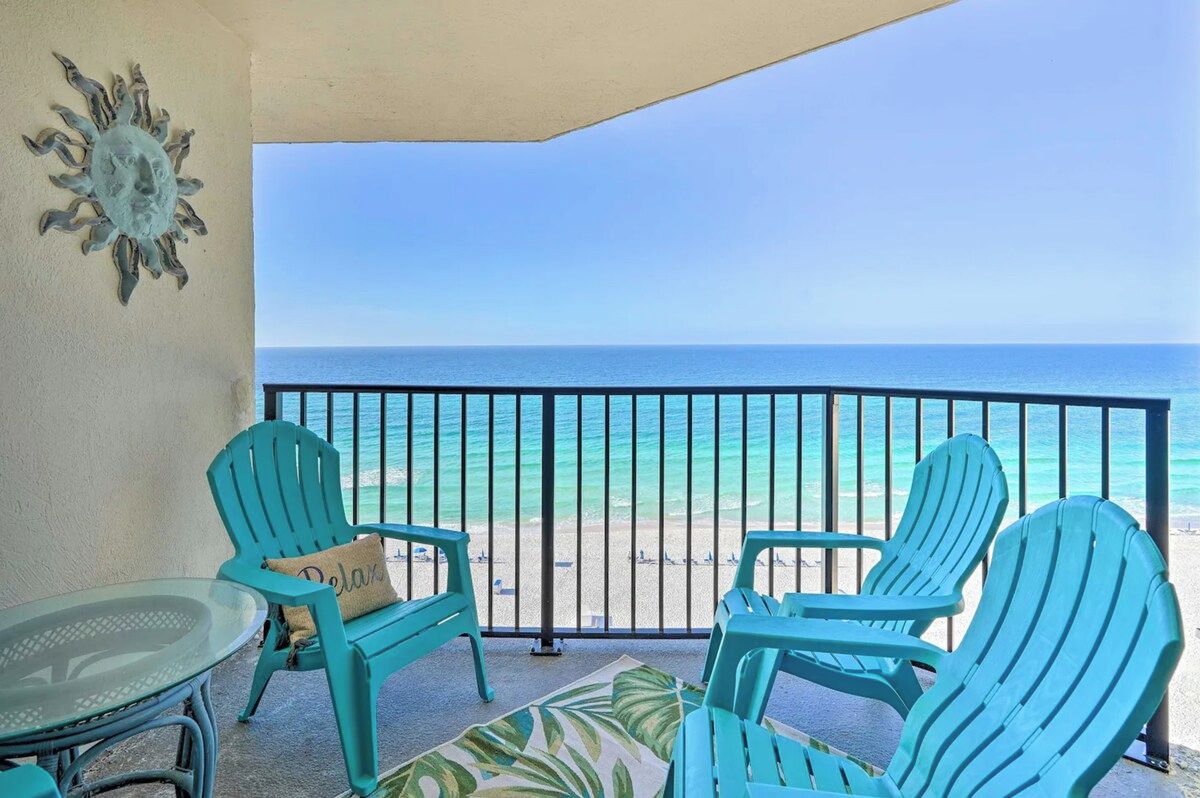 A balcony with turquoise chairs and a glass table, overlooking a beach with clear blue water.