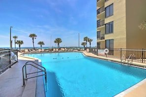 A swimming pool with a view of the ocean, surrounded by palm trees and a building with balconies.