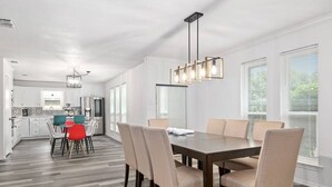 A modern kitchen with white cabinets, a dining area with a wooden table and beige chairs, and a pendant light fixture. - Stylish 5BR 3BA Home w/Pool Perfect for 16ppl (Allen)