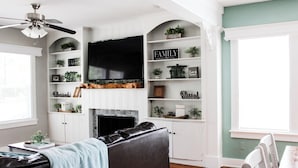 A living room with a fireplace, a TV mounted above it, and shelves with plants and family-themed decorations. - 3 Bedroom Cozy Home with the Perfect Patio (Winter Haven)