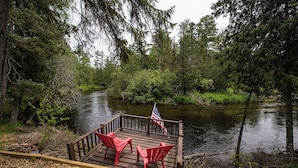 A wooden deck with two red Adirondack chairs and an American flag, overlooking a river surrounded by dense forest. - Cozy Riverside Getaway Pine Bend Cottage (Grayling)