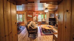 A cozy cabin interior with a wooden floor, a sofa, a dining table, a television, and a fireplace. - Cozy Riverside Getaway Pine Bend Cottage (Grayling)
