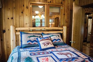 A wooden cabin interior with a bed featuring a quilt with animal prints and a wooden headboard. - Cozy Riverside Getaway Pine Bend Cottage (Grayling)
