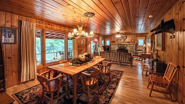 A wooden cabin interior with a dining table, chairs, a fireplace, and a television. - Cozy Riverside Getaway Pine Bend Cottage (Grayling)
