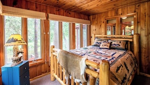 A wooden cabin bedroom with a bed, a dresser, and a window with blinds. - Cozy Riverside Getaway Pine Bend Cottage (Grayling)