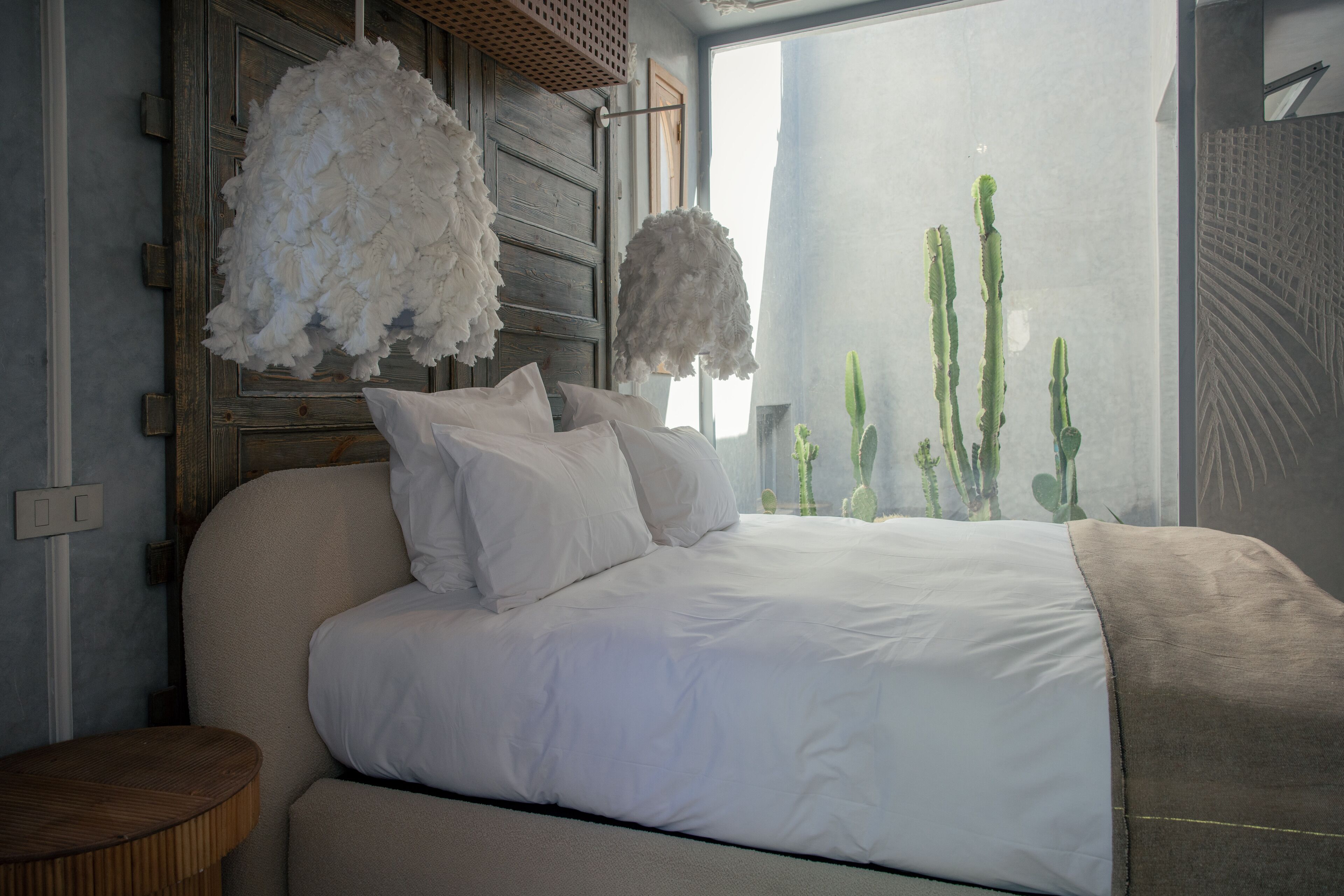 Une chambre à coucher avec un grand lit, du linge de lit blanc, une tête de lit en bois et une vue sur des cactus par la fenêtre.