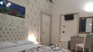 Habitación de hotel con cama y cabecera capitonada, colcha estampada, mesita de noche con lámpara, cómoda con espejo y televisor montado en la pared.
