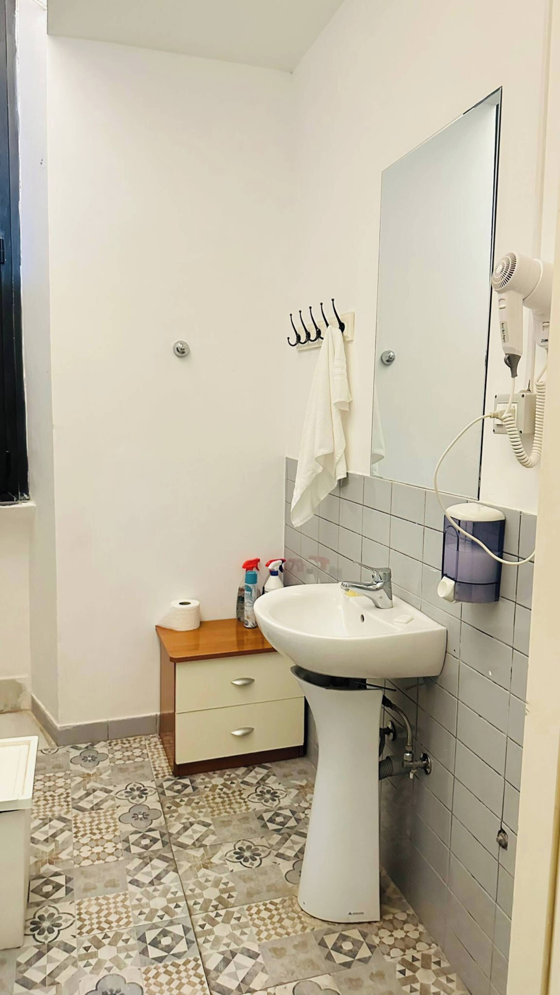 Une salle de bain avec un lavabo sur colonne blanc, un miroir, un porte-serviettes, un petit meuble en bois et un sol carrelé.