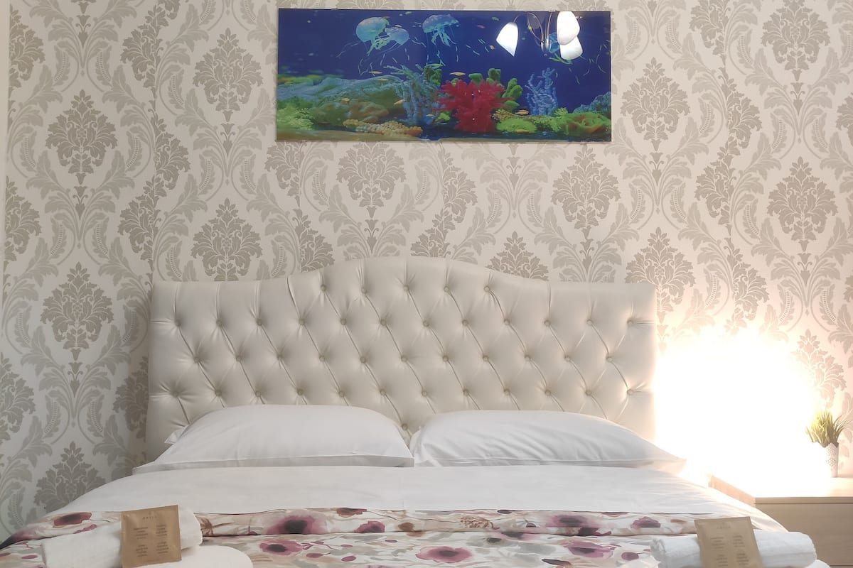 A bedroom with a tufted headboard, floral bedspread, neatly folded towels, and a framed picture of an underwater scene on the wall.