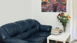 Una sala de estar con un sofá de cuero negro, una mesa de centro blanca con libros y un jarrón de flores, y un cuadro de un paisaje urbano en la pared.