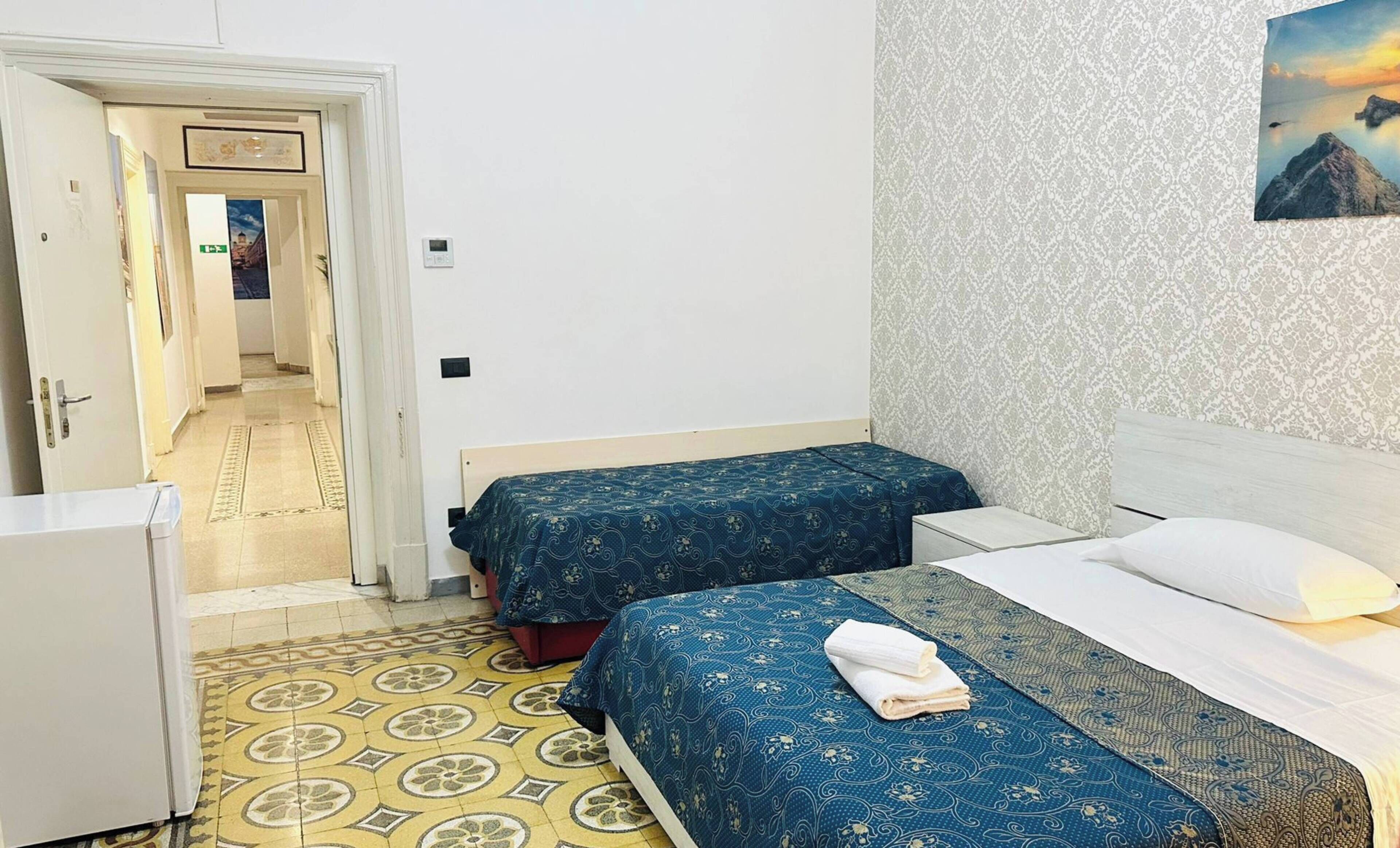 Een hotelkamer met twee bedden, een klein wit bureau, een vloer met een patroon en een schilderij aan de muur.