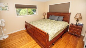 A bedroom with a wooden bed, a nightstand, a bedside lamp, a window with blinds, and a wall-mounted fan.