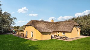 Une maison à toit de chaume, aux murs en briques, avec un petit abri attenant et une table de pique-nique dans la cour.