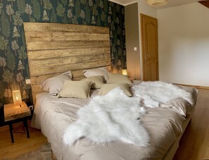 Quarto com cabeceira de madeira, cama com roupa de cama bege e almofadas brancas, mesa de cabeceira pequena com abajur e uma porta à direita.
