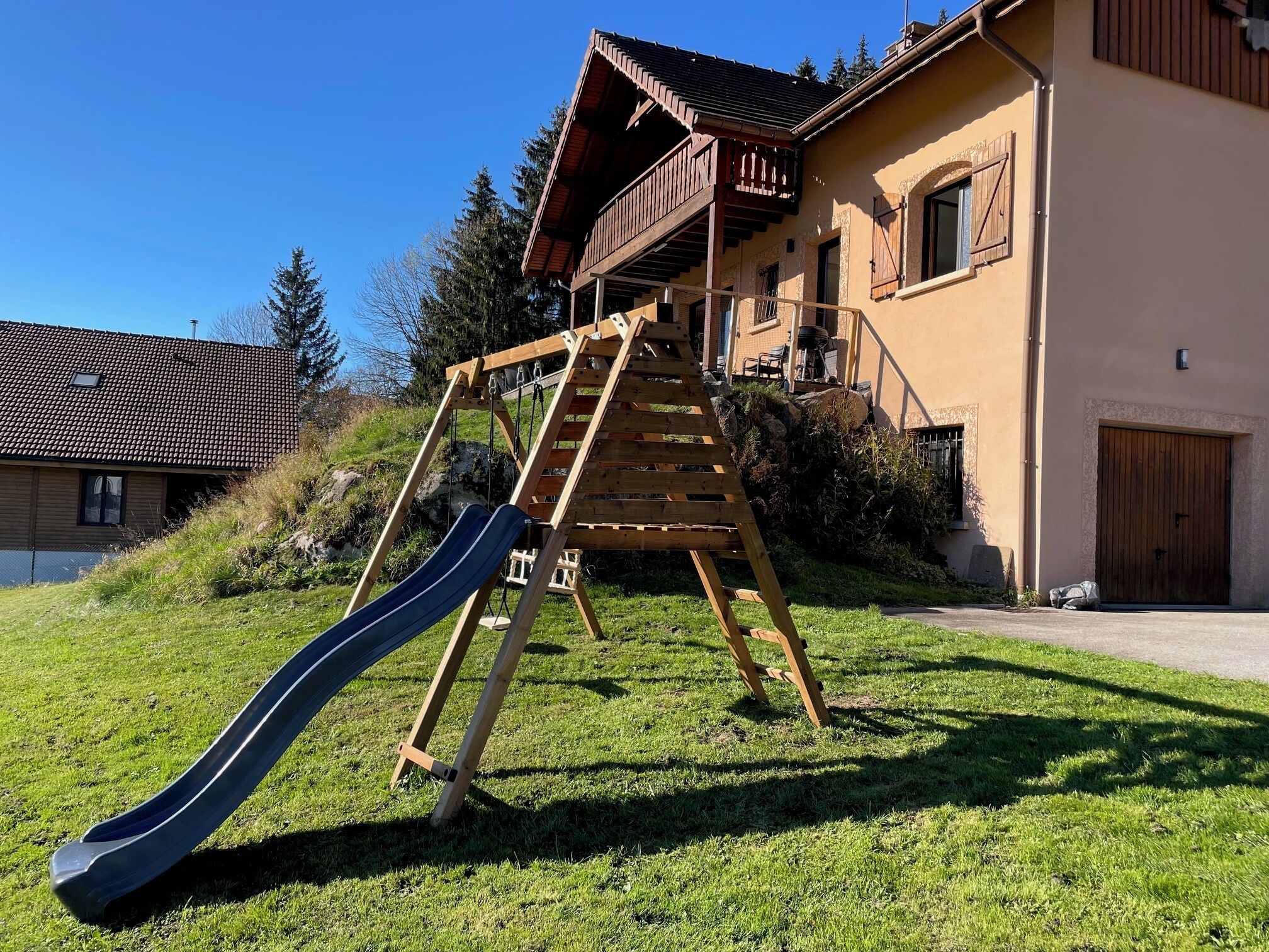 A wooden playground with a slide and a climbing structure in front of a house with a balcony.