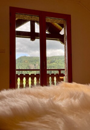 Quarto com uma grande janela, varanda de madeira e uma cama macia e branca.