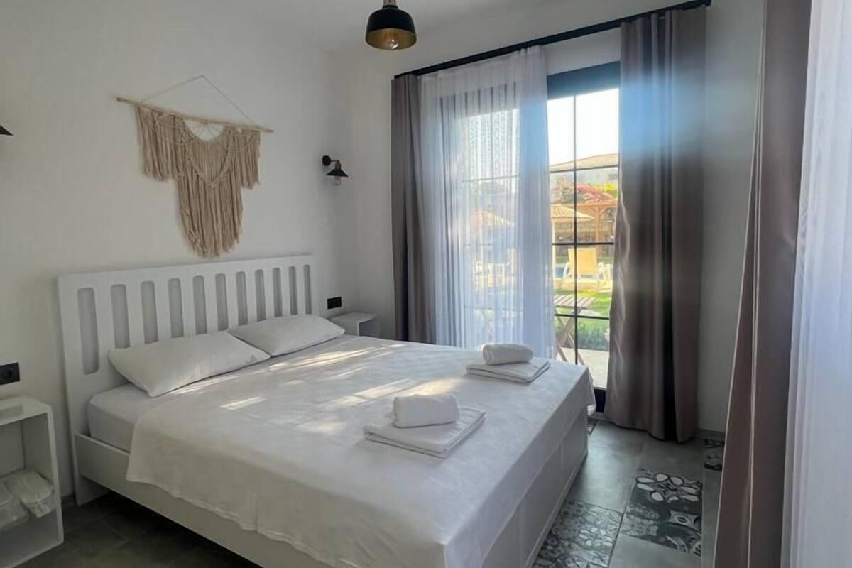 A modern bedroom with a large bed, a bedside table, a hanging macrame wall decoration, a pendant light, and a view of the outdoors through a window with curtains.