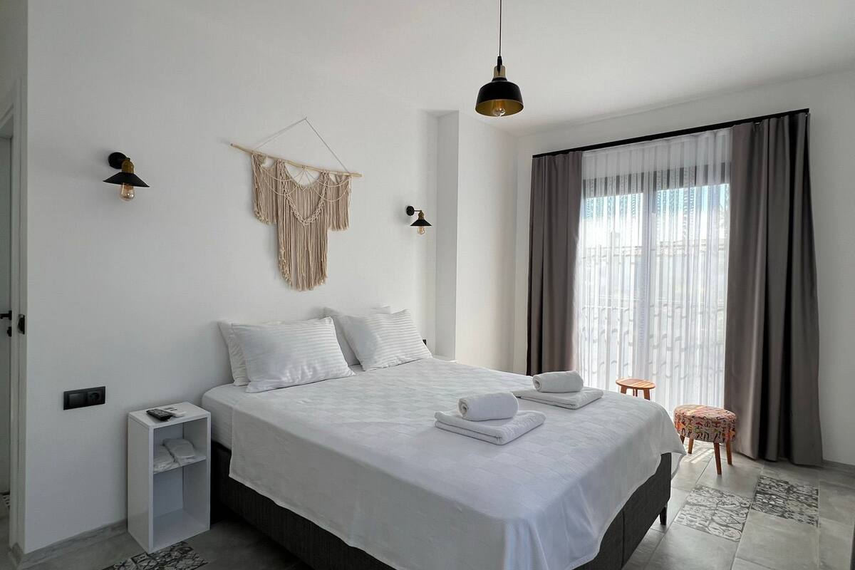 A modern hotel room with a large bed, a small bedside table, a wall-mounted macrame piece, and a window with curtains.