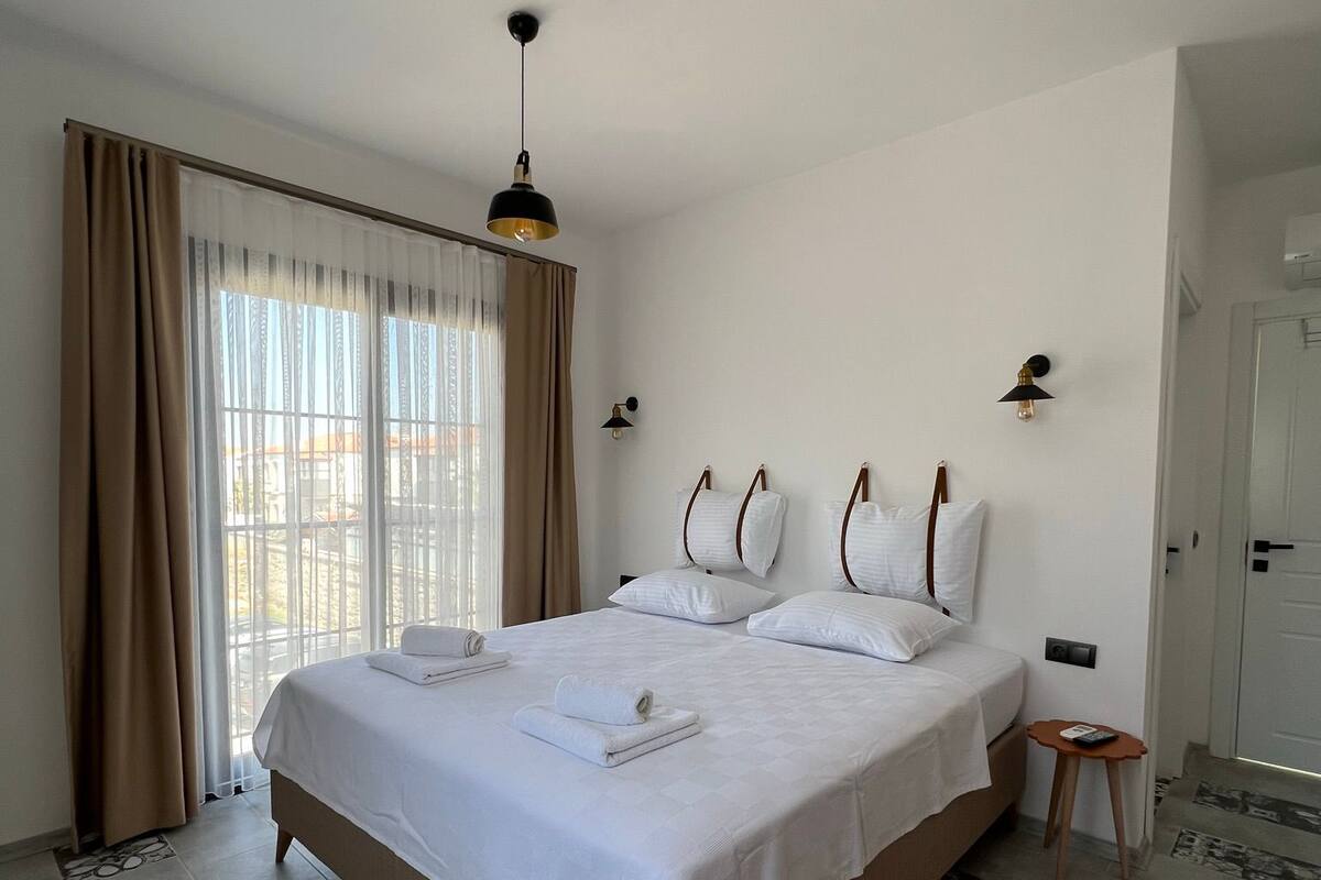 A hotel room with a large bed, a small bedside table, a nightstand, a window with curtains, and a door.