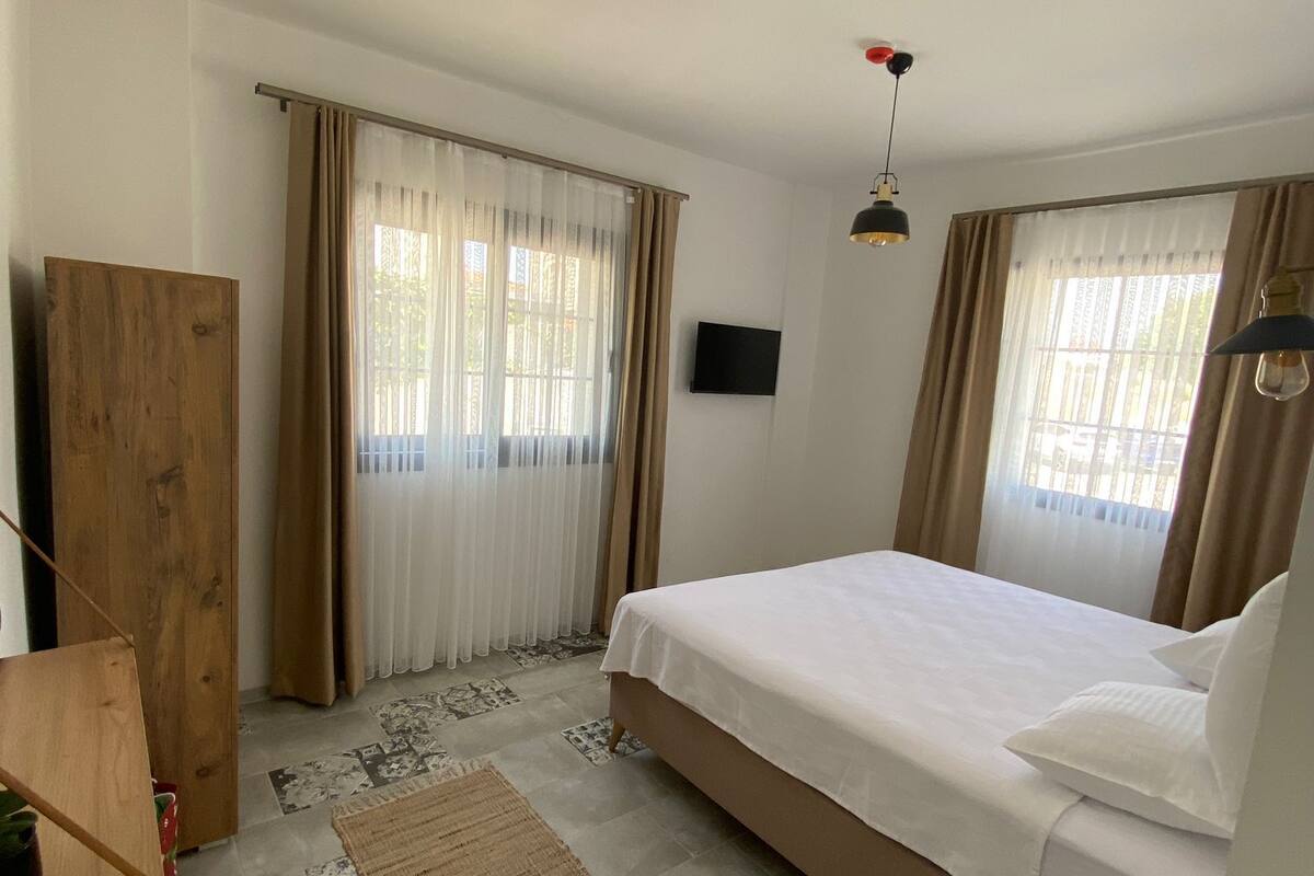 A bedroom with a bed, curtains, a television, a wooden wardrobe, a rug, and a small table with a remote control.