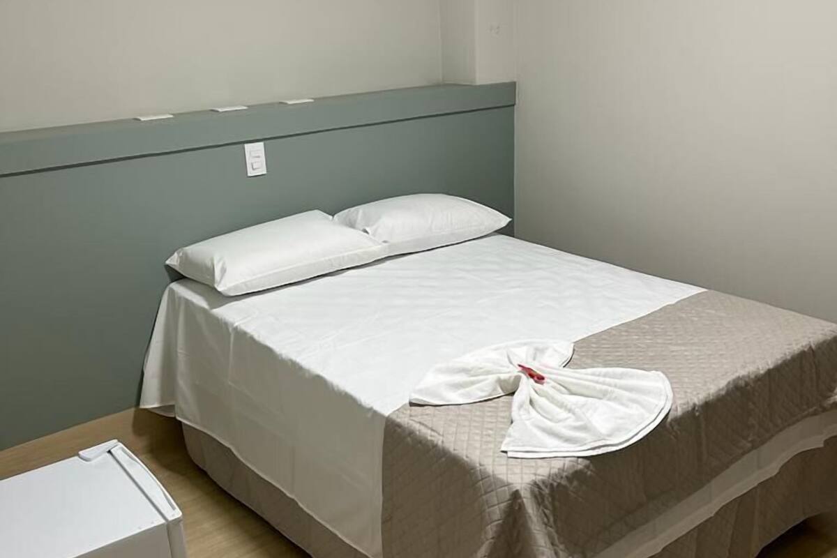 A single bed with a white bedspread and pillows, a grey headboard, and a small white appliance on the floor.