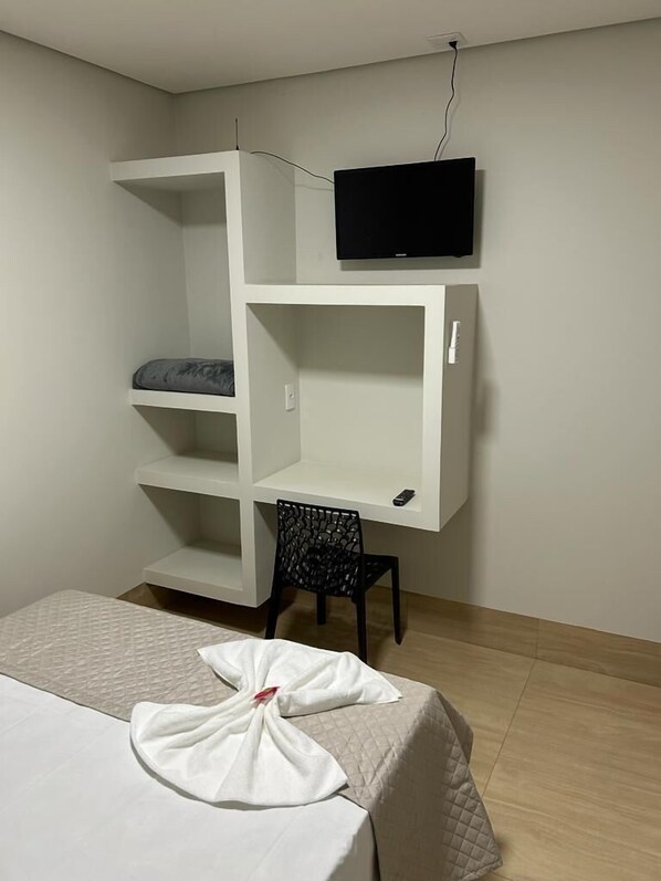 A room with a bed, a desk with a chair, and a TV mounted on the wall. - Urupês (Urupês)