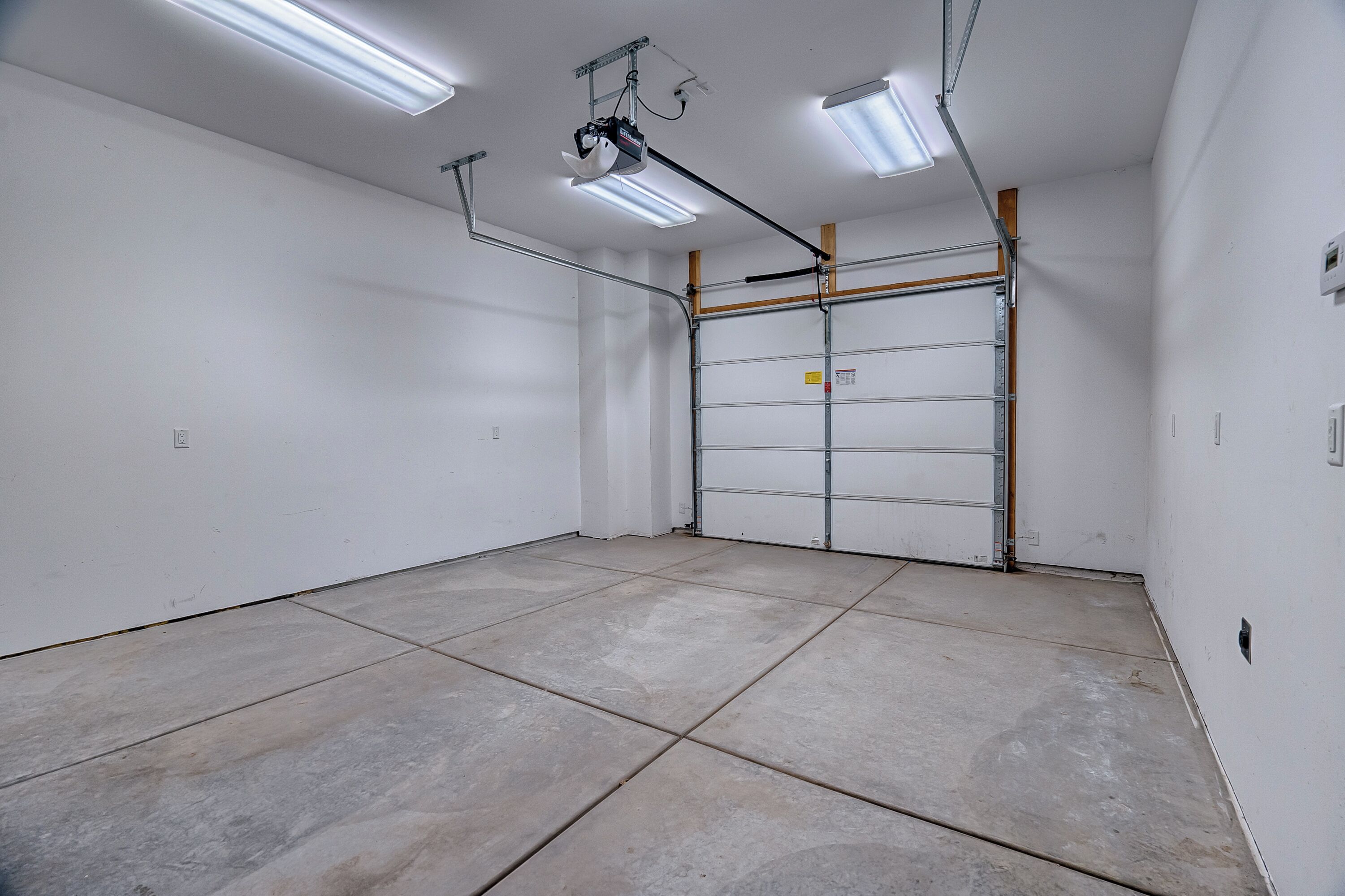 A clean, empty garage with a metal door, fluorescent lighting, and a ceiling-mounted projector.