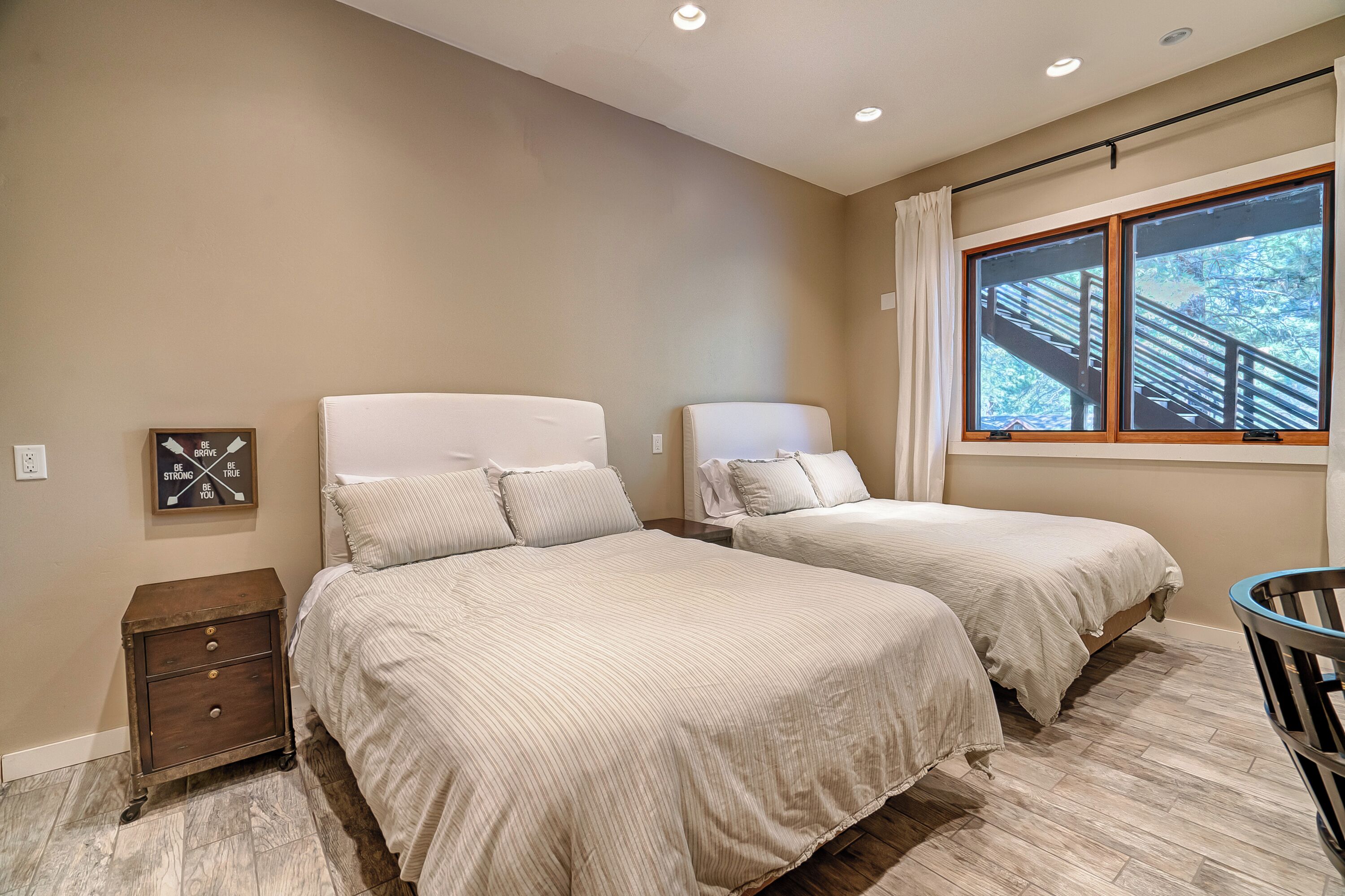 A bedroom with two beds, a nightstand, a window with a view of a staircase, and a motivational quote on the wall.