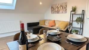 A dining table set for four with wine glasses, plates, and cutlery. A bottle of wine is on the table. The room has a sofa with cushions, a plant, and a painting on the wall. - Stylish & Spacious Apartment (Tunbridge Wells)