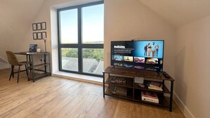 A modern living room with a large window, a wooden floor, a TV on a wooden stand, and a desk with a chair. - Stylish & Spacious Apartment (Tunbridge Wells)