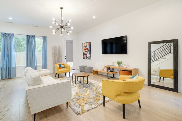 A modern living room with a sofa, armchairs, a coffee table, and a TV. There is a large mirror and a staircase visible in the background. - Modern Houston Home w/ Yard, 3 Mi to Toyota Center (Houston)